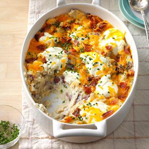 Casserole topped with cheese, bacon, and chives, partially served; sits in a white dish on a tablecloth, near teal plate and silverware.