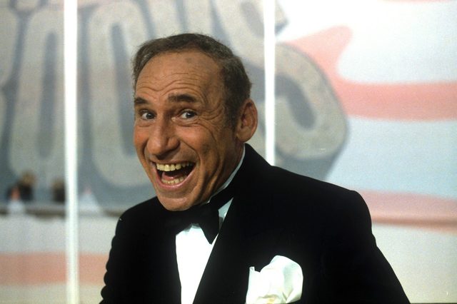Man grinning broadly, wearing a tuxedo with a white pocket square, against a colorful, blurred backdrop.