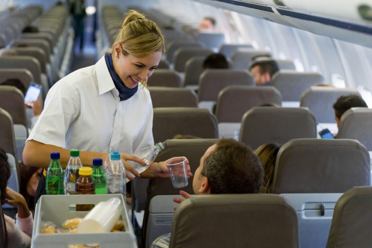 Flight attendant serves water to a passenger in an airplane aisle, with other passengers seated and occupied in a modern cabin environment.