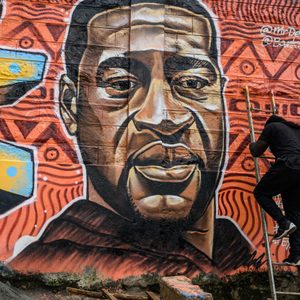 Mural on a wall depicts a large portrait; an artist climbs a ladder, surrounded by bright geometric patterns and social justice messages.