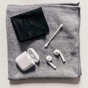 Earbuds sit with an open case, cleaning cloth, and cotton swab on a textured gray cloth background.