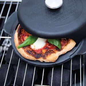 Pizza bakes under a black cooking dome on a grill with charcoal, adorned with basil leaves and melted cheese.