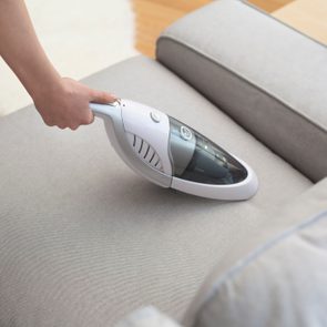 Handheld vacuum cleaning gray sofa cushion, with a potted plant and hardwood floor nearby.