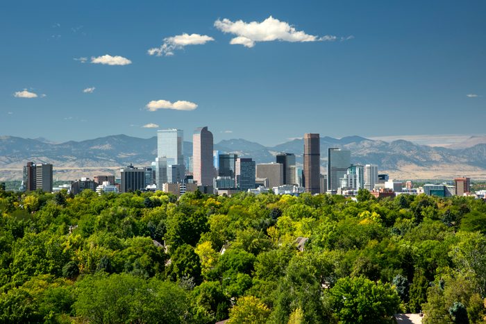Skyscrapers rise among a cityscape, framed by green trees and distant mountains under a clear blue sky with scattered clouds.