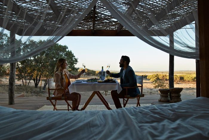 A couple dines and toasts at a table under a canopy, overlooking a scenic outdoor landscape with trees and open fields.